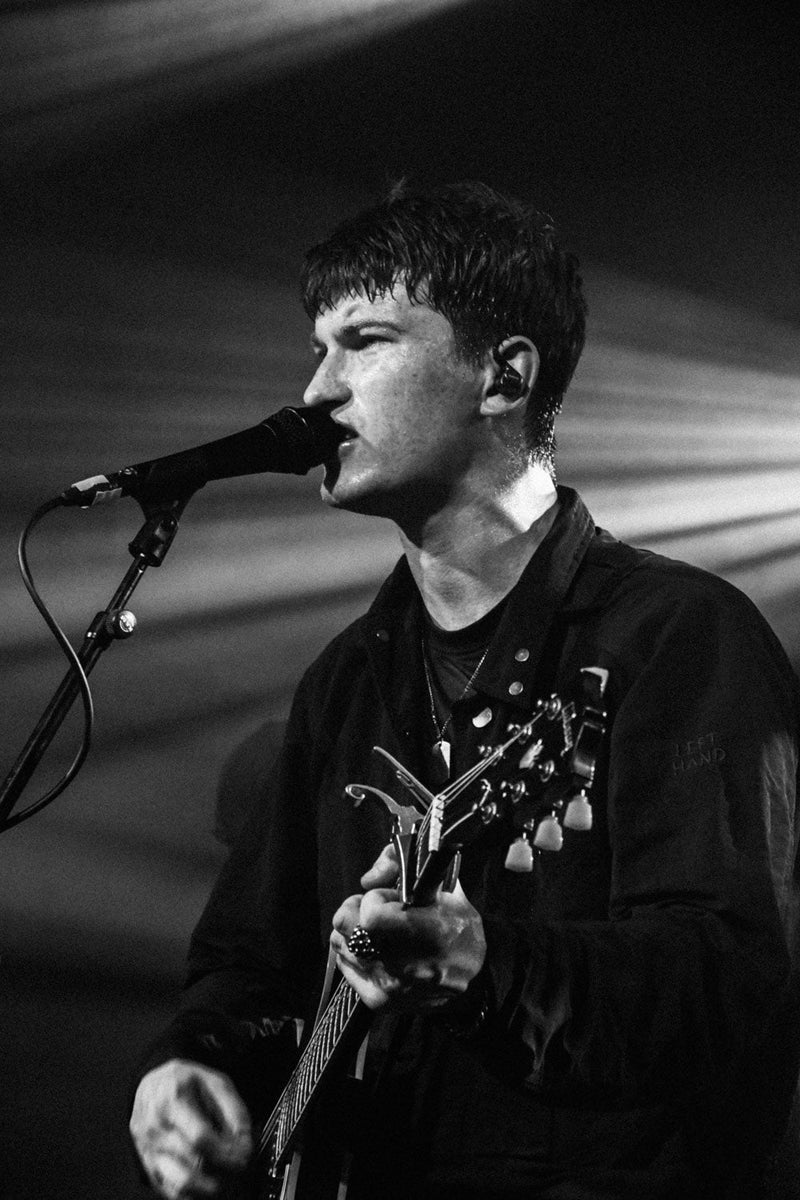 Andrew Cushin performing live on stage, singing into a microphone and playing guitar under dramatic lighting.