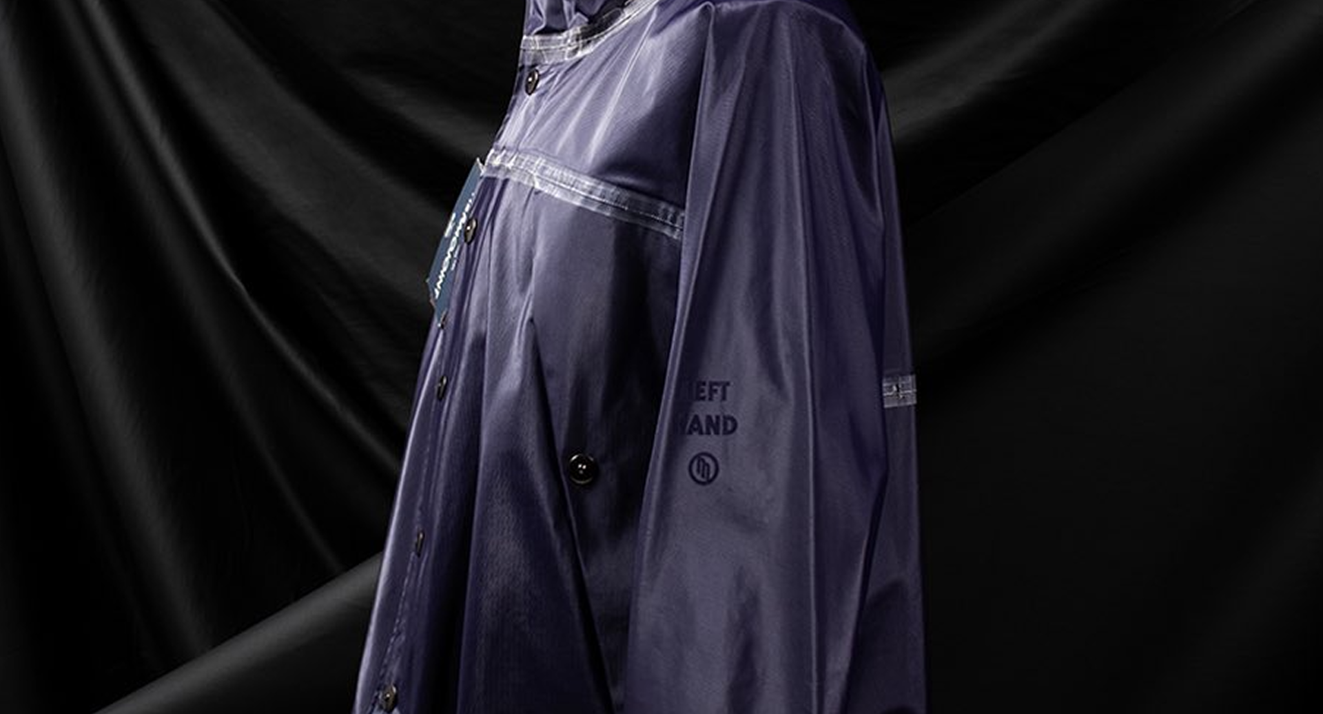 EXPLORE OUR ARCHIVE PIECES Closeup of Left Hand Jacket: dark purple waterproof design with "LEFT HAND" text, logo, buttons, and reflective trim on black backdrop.