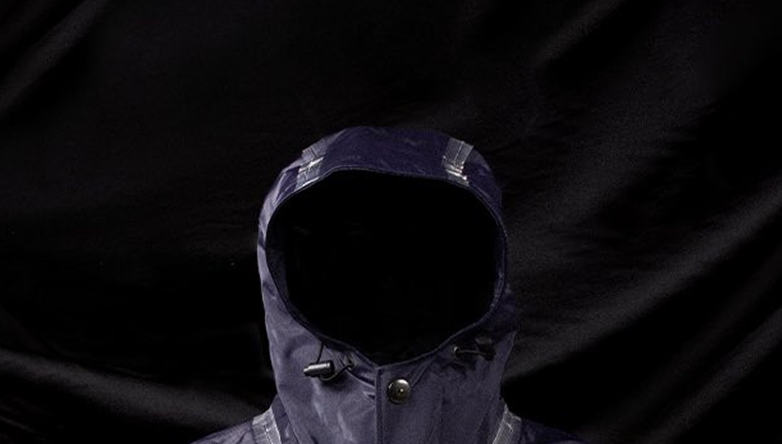 THE HISTORY Close-up of a navy Left Hand jacket with raised hood, showcasing technical fabric and taped seam detailing against a dark background.