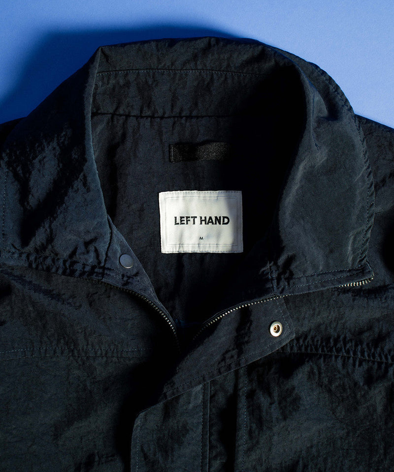 Close-up of Left Hand’s Ticino Field Jacket in navy, showing collar, zip fastening and inner label, laid flat on a blue background.