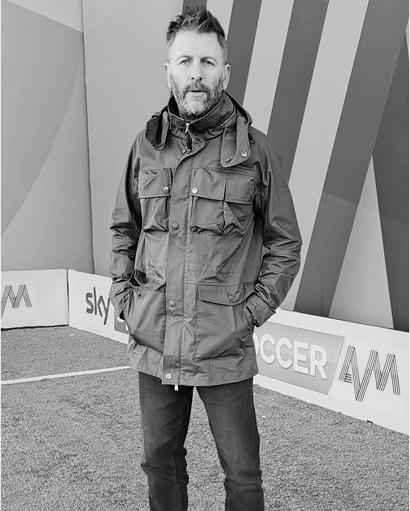 John "Fenners" Fendley standing outdoors in a casual jacket and jeans, captured in a black and white photograph.